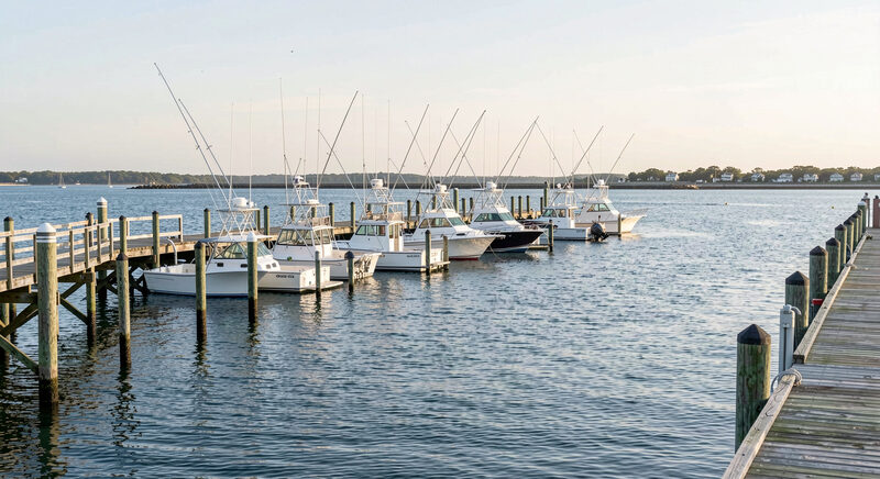 A row of sportfishing charter boats moored along a weathered wooden pier in a small Cape Cod Bay harbor at morning high tide