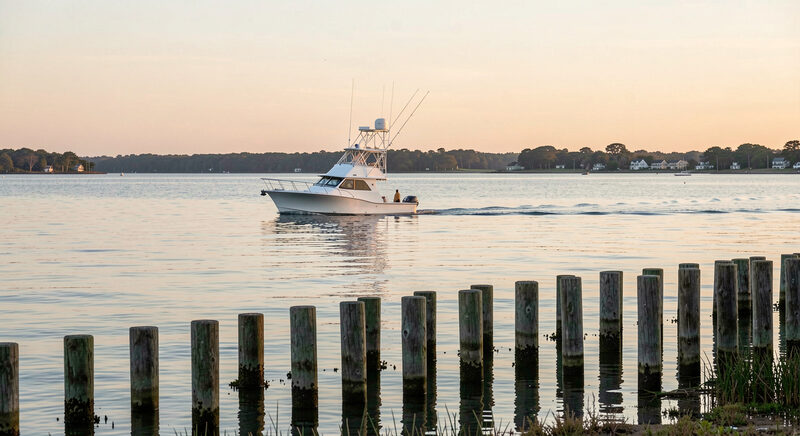 A small sportfishing boat crossing calm Cape Cod Bay at sunrise, a line of weathered wooden channel marker posts in the foreground shallows, no people.